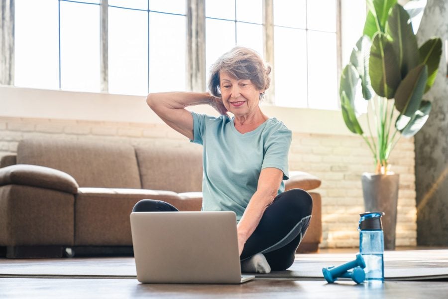 sporty grandmother doing sport on fitness mat in training outfit while using laptop for online video